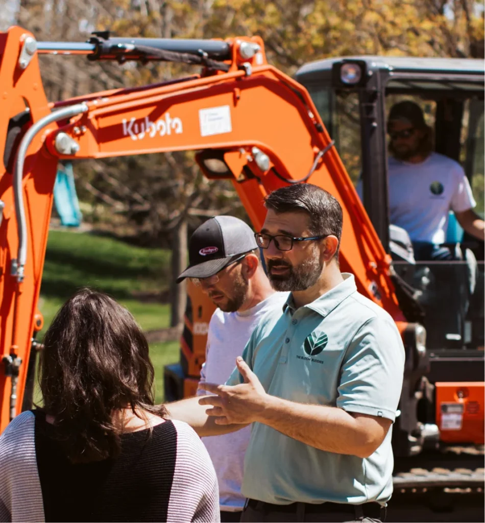 Landscape business owner discussing an active residential jobsite with a client and crew while compact equipment operates in the background.